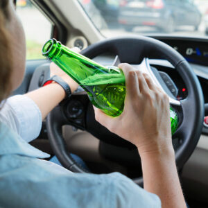 A person behind the steering wheel of a car, drinking a green bottle of beer.