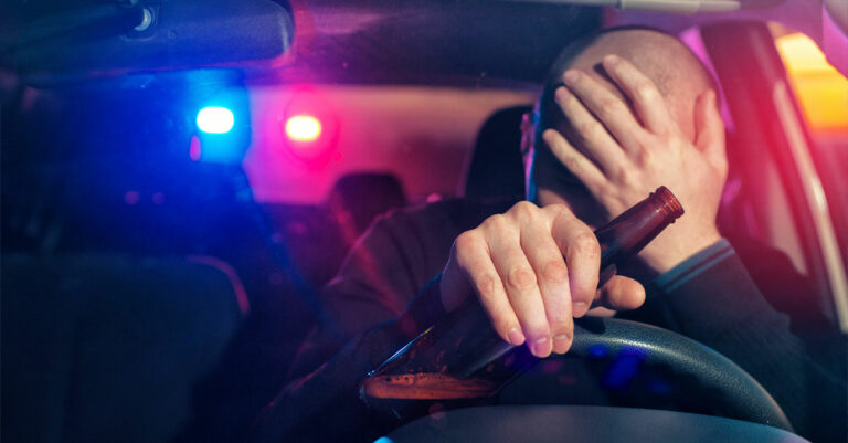 A man getting pulled over by police, with a bottle of beer in his left hand while gripping the steering wheel.